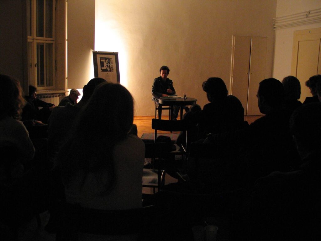 Особа седи сама за столом на осветљеној сцени, обраћајући се публици у слабо осветљеној просторији. Урамљено уметничко дело стоји близу прозора, док сенковите фигуре из контекст колектива пажљиво слушају, отелотворујући уметност и солидарност кроз активизам.