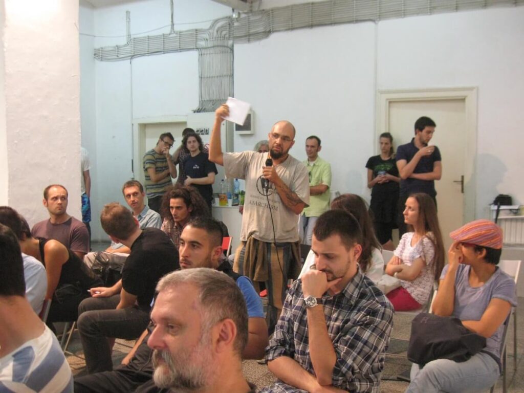 Човек стоји у препуној просторији држећи микрофон и комад папира, изгледајући као да говори или поставља питање о уметности или активизму. Људи пажљиво слушају, стварајући атмосферу колективне солидарности на овом неформалном састанку.