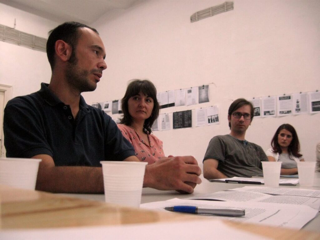 Четири особе седе за столом и разговарају, са папирима, пластичним чашама и оловком испред себе. Одштампане странице су изложене на зиду иза њих у светлој, минималистичкој просторији, одражавајући дух уметности и солидарности Контекст колектива.