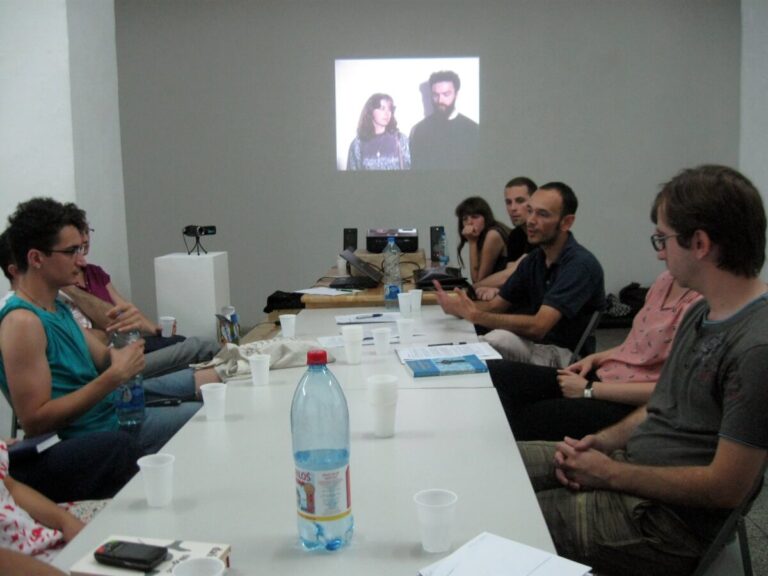 Група људи седи око стола у учионици и разговара о уметности и солидарности. Папири и флаше воде су разбацани по столу, док се на зиду иза њих појављује пројектована слика две особе.