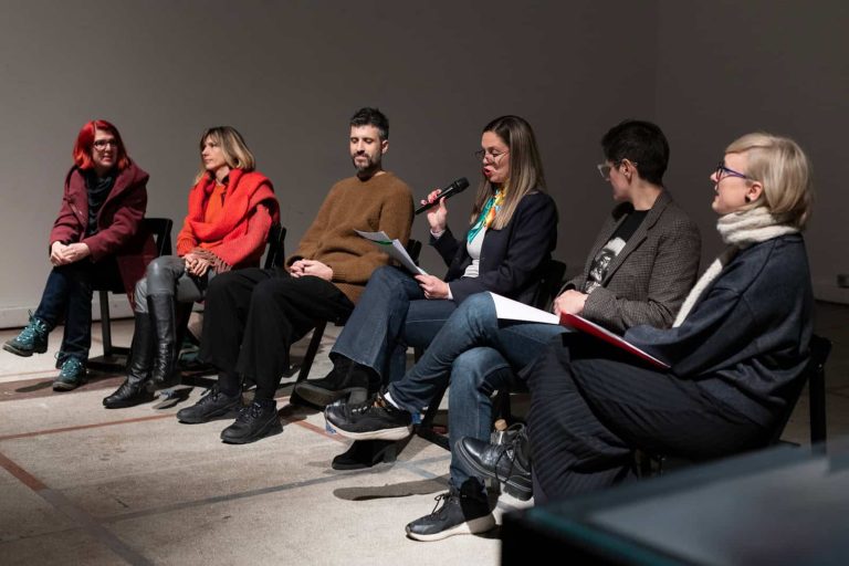 Шест људи седи у реду на столицама током панел дискусије о уметности и активизму. Једна особа држи микрофон и говори, док остали пажљиво слушају. Окружење је у затвореном простору са минималном позадином.