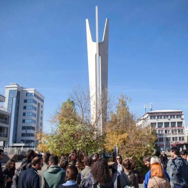 Група људи стоји испред високог, белог, трокраког бетонског споменика – симбола уметности и солидарности – окруженог дрвећем и модерним зградама под ведрим плавим небом.