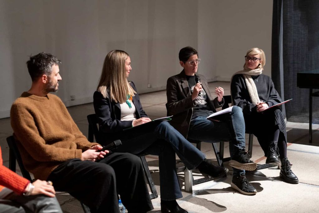 Четири особе седе на столицама у затвореном простору и учествују у панел дискусији о уметности и активизму. Једна особа говори у микрофон док остале пажљиво слушају у овом професионалном, добро осветљеном окружењу.