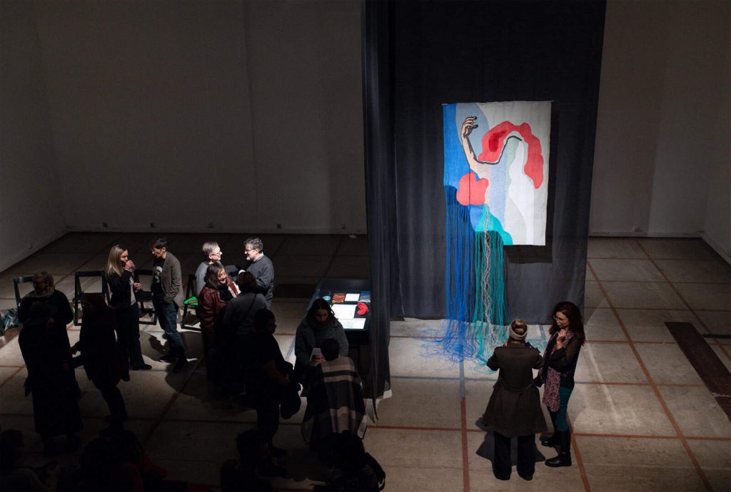 Група људи стоји и разговара у слабо осветљеном галеријском простору, делу догађаја Контекст колектив уметност, близу шареног текстилног уметничког дела које приказује апстрактну фигуру са црвеном косом и испруженом руком, која виси са црне завесе са плавим нитима.