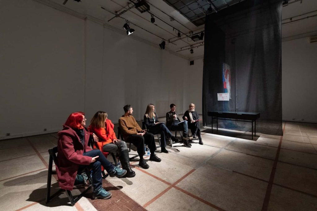 Шест људи седи у реду столица у слабо осветљеној, модерној галерији коју је курирао Контекст колектив, окренути ка црној мрежастој завеси иза које се налазе уметничка дела. Минималистички, индустријски простор одјекује темама уметности, солидарности и активизма.