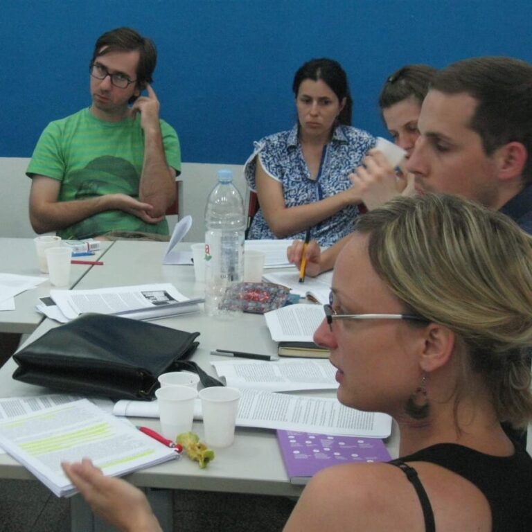 Пет људи седи око стола са папирима, свескама, шољама и флашом воде, учествујући у дискусији о колективној уметности. Једна жена говори док остали пажљиво слушају, одражавајући солидарност и колаборативни активизам.