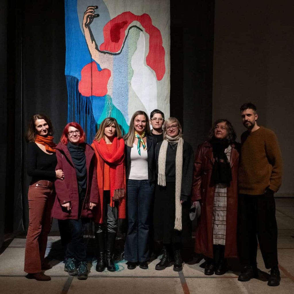 Осам људи стоји заједно у затвореном простору испред шареног, апстрактног текстилног уметничког дела са смелим црвеним, плавим и беж облицима, са фигурама подигнуте руке. Већина групе носи шалове и тамну одећу, осмехујући се у камеру.