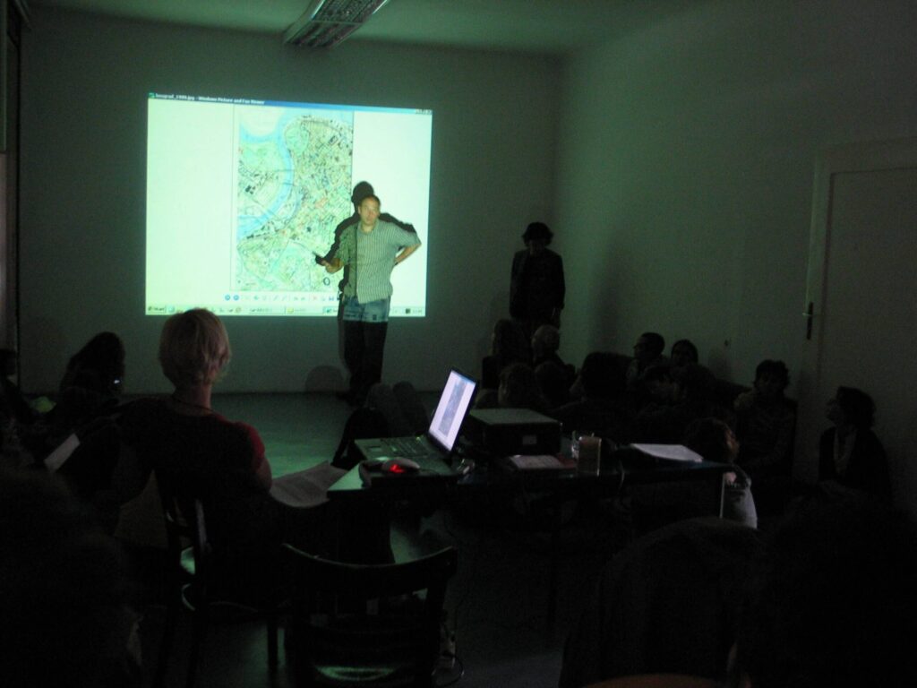 Особа стоји и говори испред пројектоване мапе, обраћајући се слабо осветљеној просторији пуној људи који седе – сцена која одражава уметност и солидарност. У првом плану је сто са лаптопом и другом опремом.