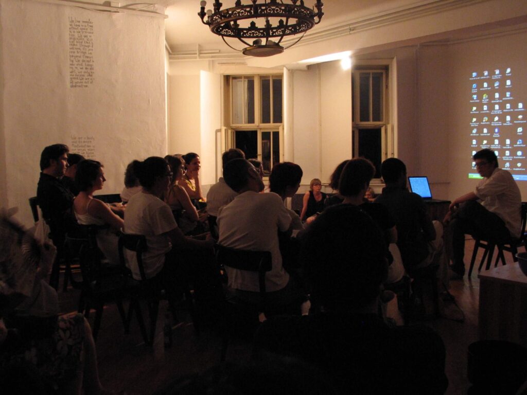 Група из Контекст колектива седи у слабо осветљеној соби, окренута ка пројектованој рачунарској површини, док једна особа презентује поред лаптопа — подстичући уметност, солидарност и активизам кроз заједничку дискусију.