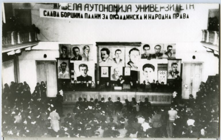 Велика група се окупља у сали где су портрети и фотографије изложени на сцени, испод ћириличног транспарента. Публика пажљиво слуша док говорници говоре о уметности, солидарности и колективном активизму.