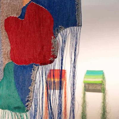 Крупни план шареног тканог текстила Контекст колектива са лабавим плавим нитима, са апстрактним облицима у црвеној, зеленој и плавој боји. У замућеној позадини Контекст галерије, две шарене коцке са нитима висе испод њих.