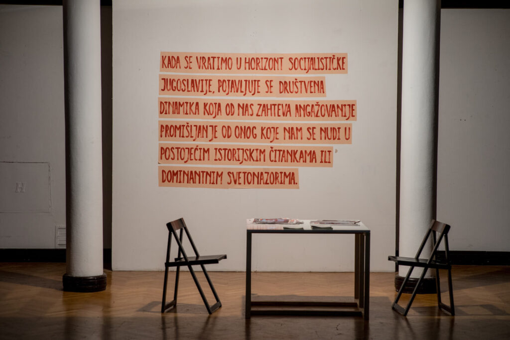 Две празне столице окренуте су ка малом столу са књигама и папирима, што евоцира уметност и солидарност. Руком писана порука на српском језику је залепљена на зид између два висока бела стуба, наговештавајући дух активизма Контекст колектива.