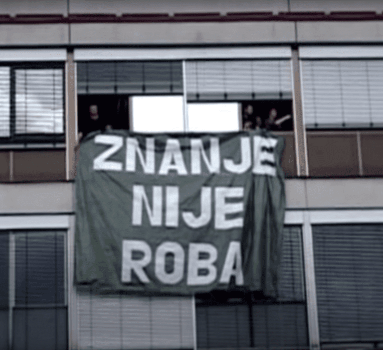 Велики зелени транспарент са белим текстом на којем пише ЗНАЊЕ НИЈЕ РОБА виси са прозора зграде, иза којег стоје чланови Контекст колектива, истичући уметност и солидарност кроз свој активизам.