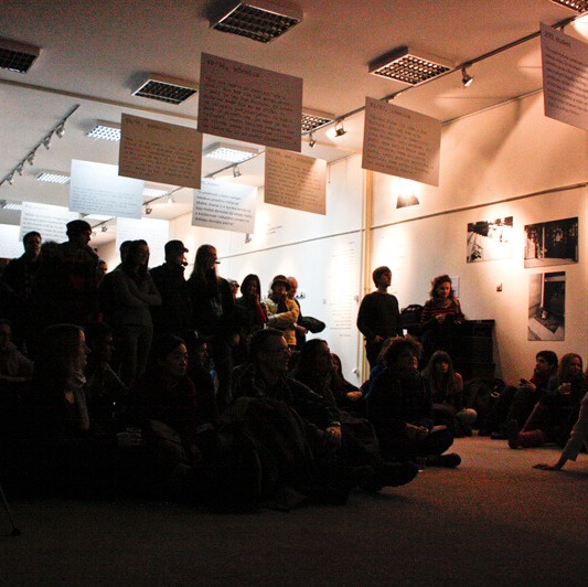 Група људи седи и стоји близу једни другима у слабо осветљеној просторији, окренути према напред. Велики листови папира са рукописом писаним текстом висе са плафона изнад њих, одражавајући дух колективне уметности и солидарности кроз активизам.