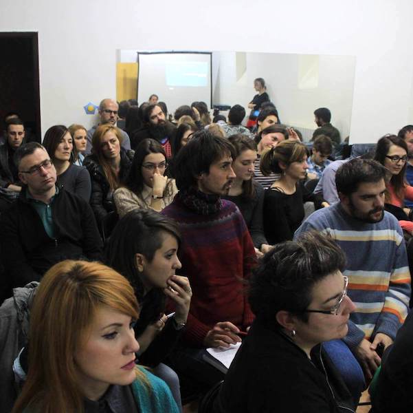 Група људи седи близу једни других у просторији, пажљиво пратећи презентацију о колективној уметности и солидарности пројектовану на платну. Већина делује фокусирано, неки праве белешке, а презентер је видљив близу платна.