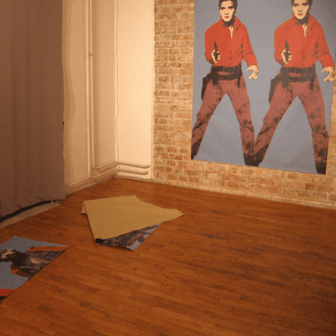 Велика фотографија Контекст колектива, на којој је мушкарац у црвеној кошуљи са пиштољима, постављена је на зид од цигле; два слична отиска делимично су уролана на дрвеном поду, истичући уметност и солидарност у иначе празној просторији са голим зидовима и завесом.