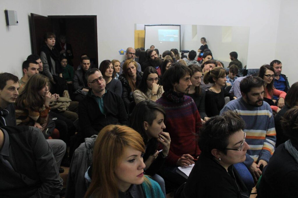 Група људи, и мушкараца и жена, седи близу један другом у просторији окренутих напред, делујући пажљиво и фокусирано. У овом контекст колективном окружењу, пројектор приказује слику на платну у позадини.