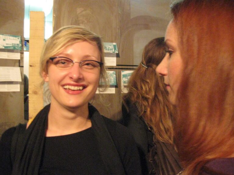 Две жене стоје у затвореном простору на догађају Контекст колектива — једна се смеши камери са наочарима и црном мајицом, док је друга гледа из профила. Замагљени људи и слике на зиду наговештавају уметност, солидарност и активизам у позадини.