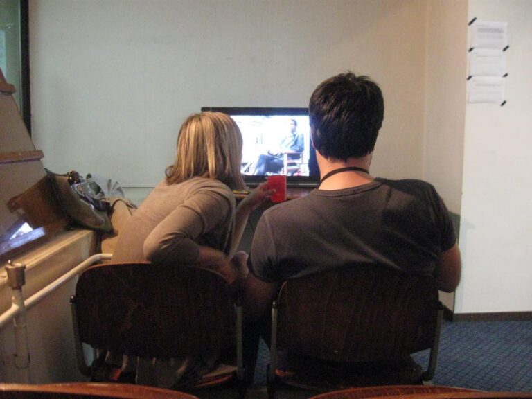 Двоје људи седе близу једно другом на дрвеним столицама, гледајући програм на малом телевизору. Једна држи црвену шољу. Неформална соба наговештава колективну енергију - интиман простор за уметност и свакодневну солидарност.