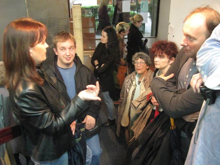 Група људи стоји близу једна другој у затвореном простору, заокупљена разговором. У овом контексту колектив, жена у црној јакни гестикулира док други пажљиво слушају, одражавајући дух солидарности и радозналости. Видљиви су капути и торбе.