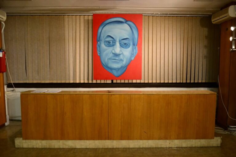 Велики, строги портрет старијег мушкарца са наочарима у плавим тоновима – симбол уметности и активизма – виси на црвеној позадини изнад дрвеног пулта у соби са беж вертикалним ролетнама и пригушеним осветљењем.