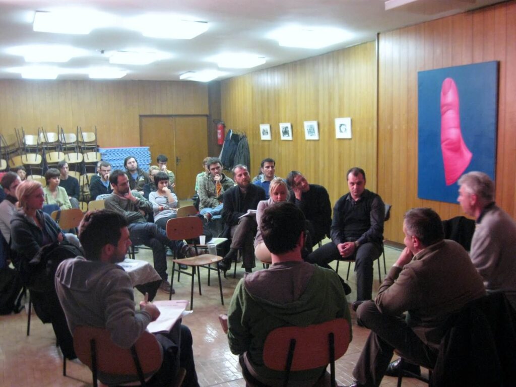 Група људи седи у кругу и разговара о уметности и солидарности у просторији са зидовима обложеним дрветом. Празне столице су наслагане у позадини, а на зиду виси велика слика ружичастог прста.