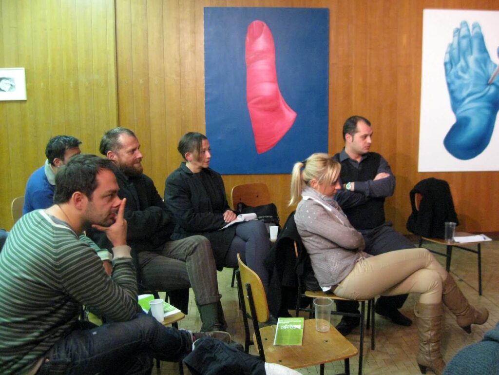 Група људи пажљиво седи у учионици са дрвеним зидовима, учествујући у контексту колективне уметности. Велике шарене слике црвеног палца и плаве руке висе иза њих, док разговарају о солидарности и активизму са свескама и пластичним чашама у рукама.