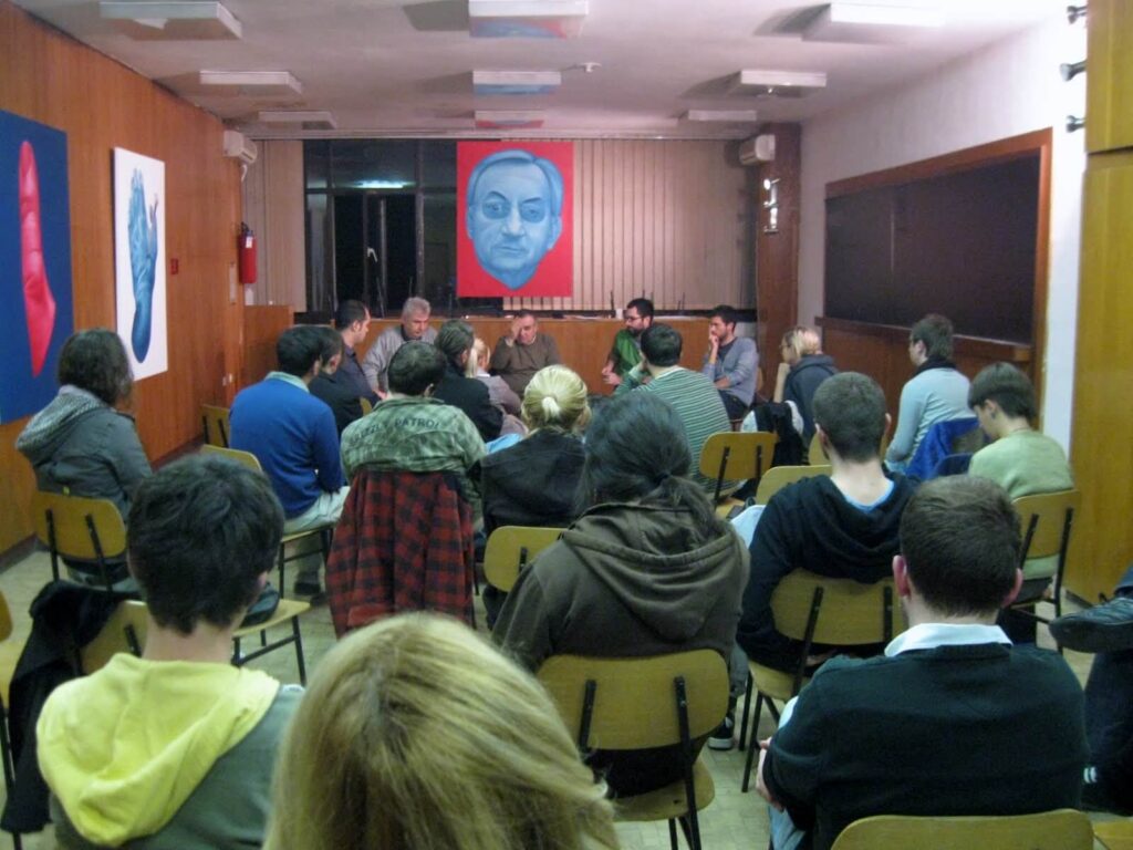 Група људи седи на столицама окренута ка панелу у предњем делу собе, учествујући у дискусији о уметности и активизму. Шарени портрети висе на зидовима обложеним дрветом иза учесника панела.