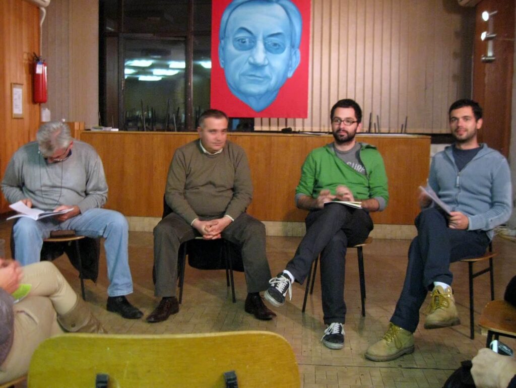 Четири мушкарца седе у полукругу на столицама у соби обложеној дрветом, разговарајући о уметности и држећи папире. Велики портрет мушког лица у плавим тоновима, који евоцира колективност и солидарност, виси на зиду иза њих.