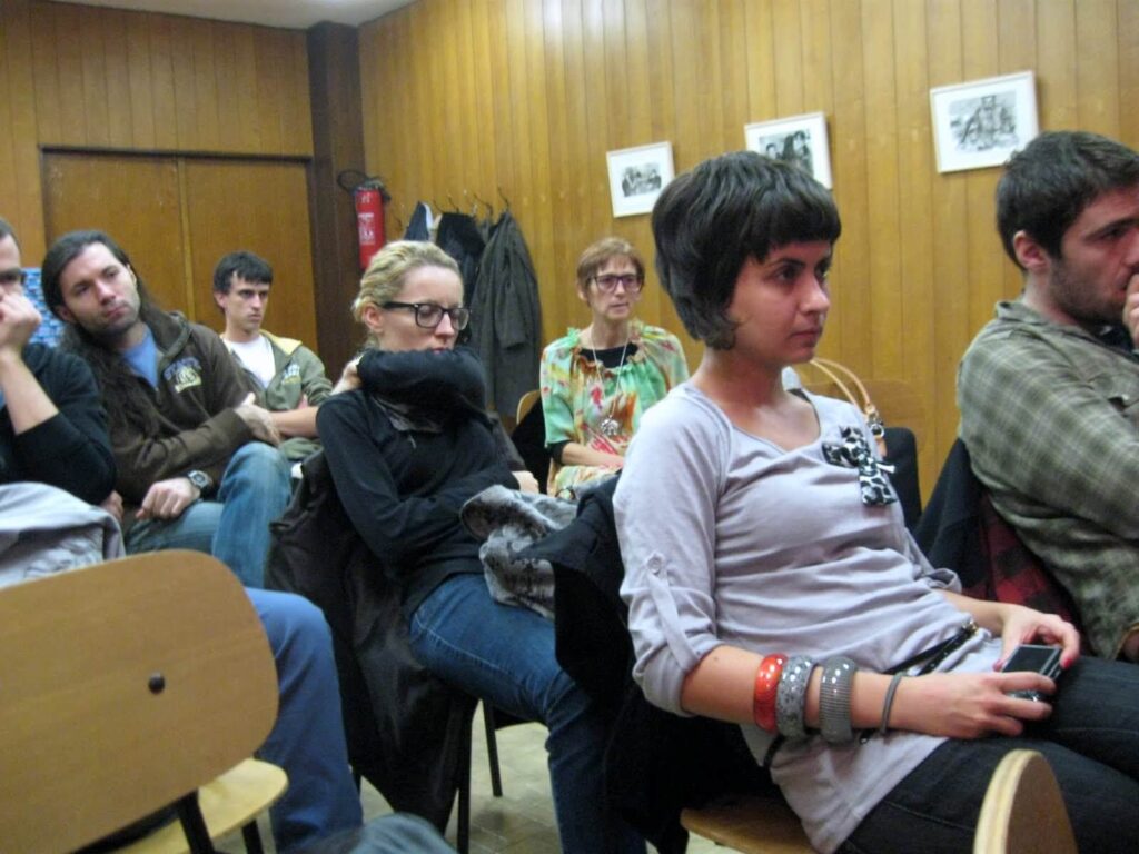 Група људи пажљиво седи у соби обложеној дрветом, отелотворујући колективност и солидарност, док урамљене слике висе на зиду иза њих. Већина изгледа пажљиво слуша, заокупљена тренутком обликованим уметношћу и активизмом.