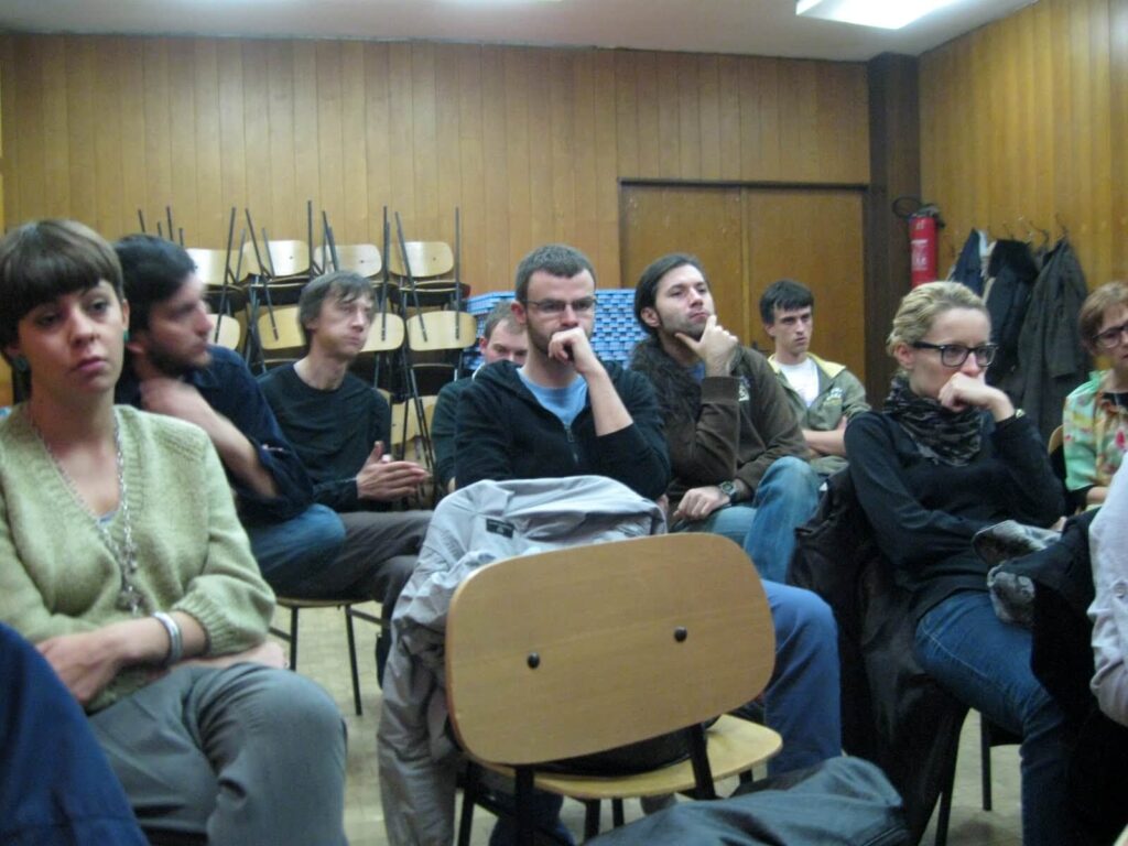 Група људи пажљиво седи у просторији са дрвеним зидовима, њихови изрази лица одражавају промишљену ангажованост. Атмосфера сугерише колектив усмерен на уметност или активизам, док наслагане столице наговештавају окупљање утемељено у солидарности.