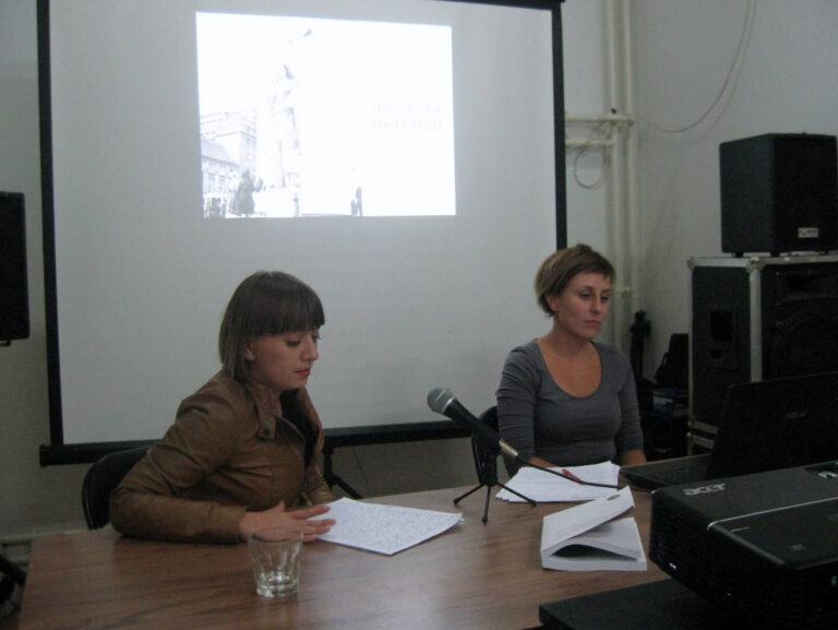 Две жене из Контекст колектива седе за столом са папирима и микрофоном, испред пројектованог презентационог платна у слабо осветљеној просторији. Једна говори о уметности и активизму, док друга гледа у свој лаптоп.