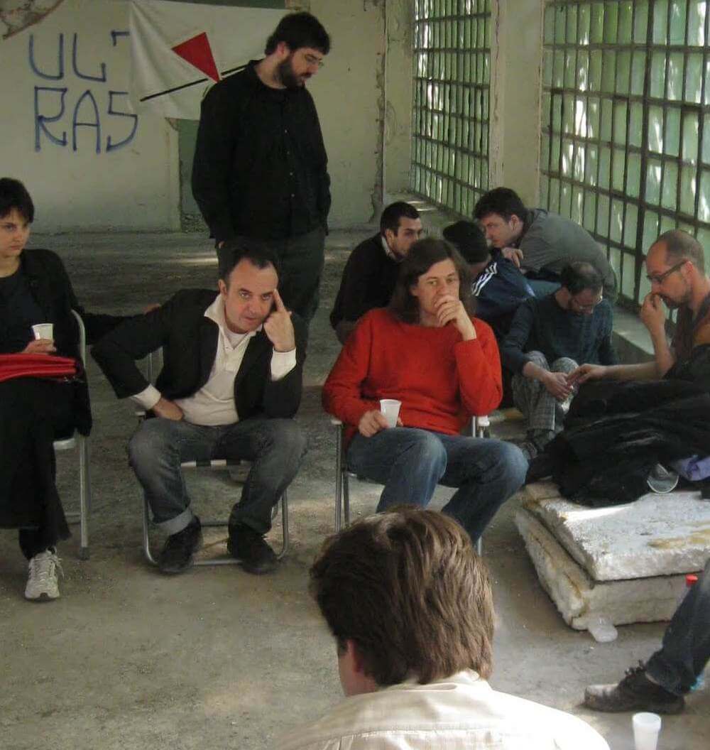 Група људи седи и стоји у истрошеној, индустријској просторији са великим стакленим прозорима. Неки разговарају, а неки ћуте. На зиду иза њих виси транспарент са натписом и црвеним троуглом - симболима колективне уметности и солидарности.